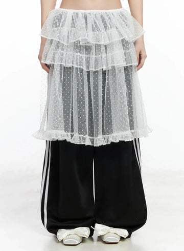 LEWKIN Tiered Tulle Polka Dot Skirt IM506 in white, front view on model, sheer polka dot tulle with multi-tier ruffles and ruffle hem, layered over black track pants with bow flats.