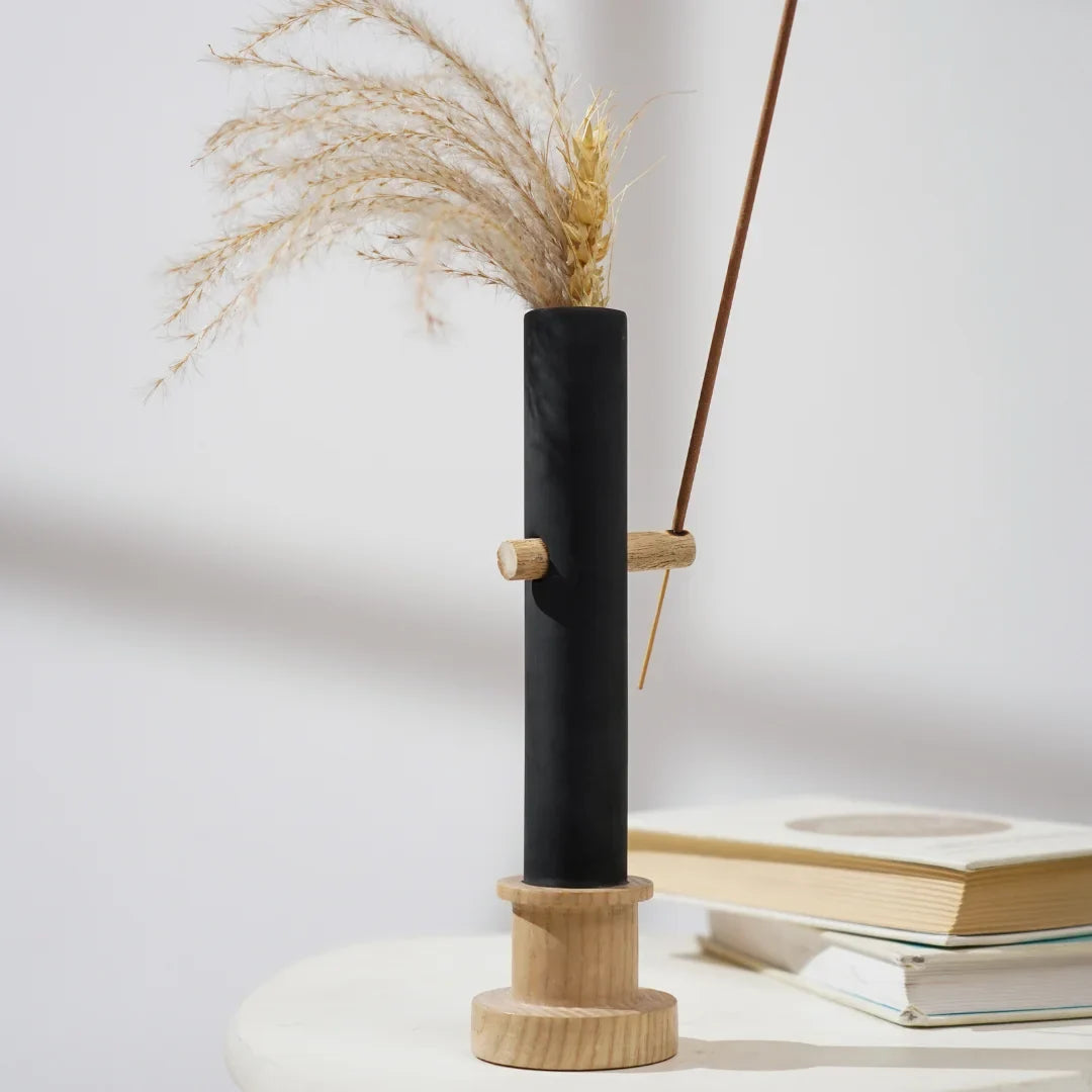 Black cylindrical vase with wooden base and top, containing dried plants on a light surface.