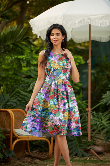Woman wearing a colorful floral dress standing outdoors with greenery and an umbrella in the background.