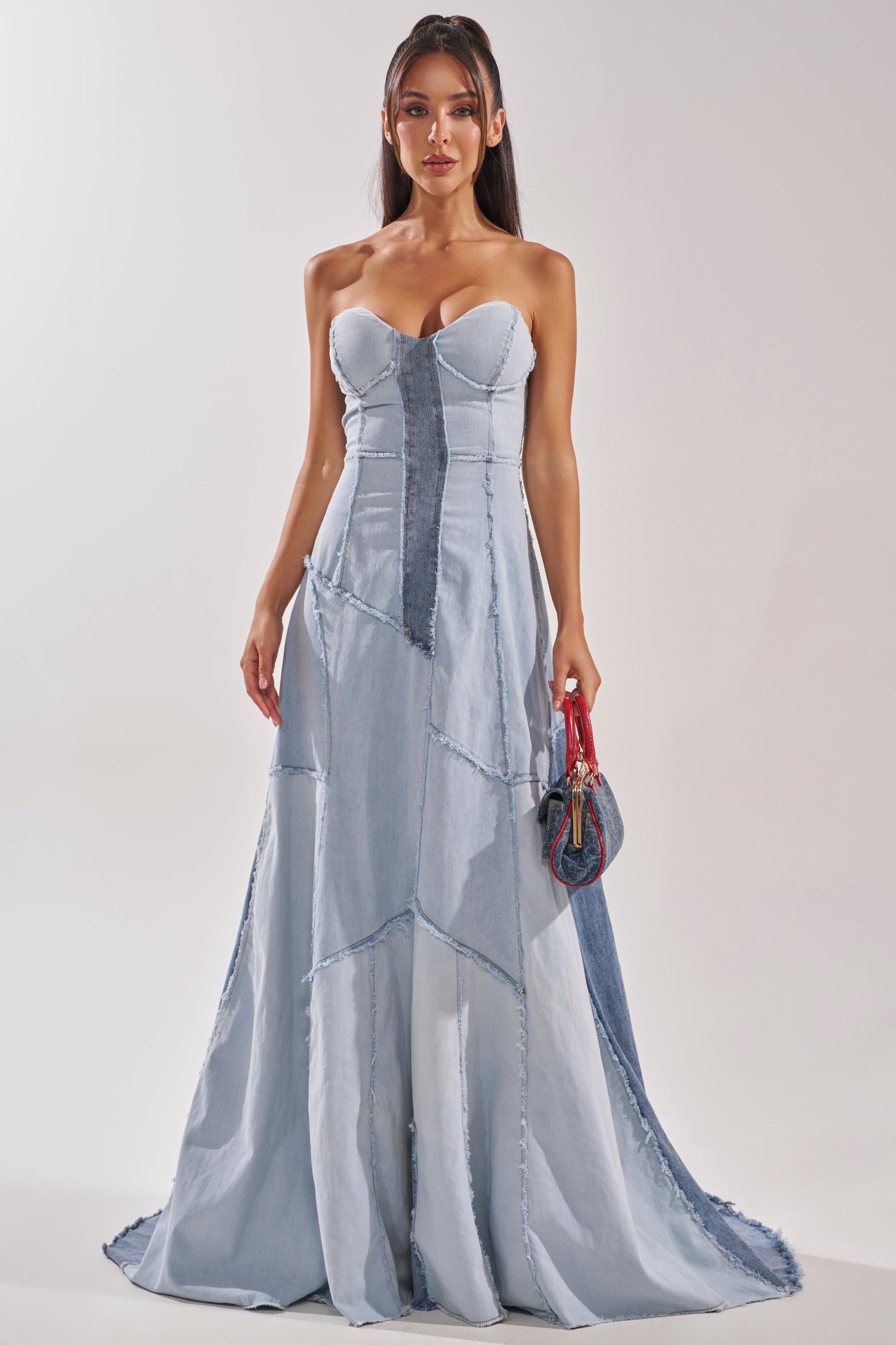 A woman wears the IT'S BRITNEY PATCHWORK DENIM MAXI DRESS, featuring strapless light and dark blue denim panels. She holds a small handbag, her long dark hair down, against a plain light-colored background.