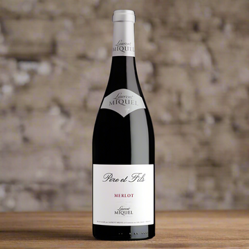 Bottle of red wine with 'Laurent Miquel' written on a white label. Merlot is written in red. The background is a brick wall and the bottle is sat on a wooden table