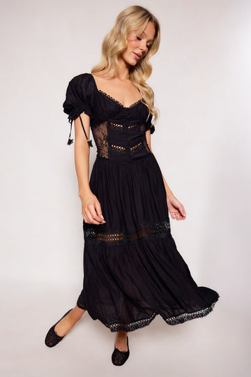 A woman with long blonde hair wears the Peregrine Dress in Black by Ivy City Co, featuring lace details and puffed sleeves. She stands against a plain light background, smiling softly while looking down.