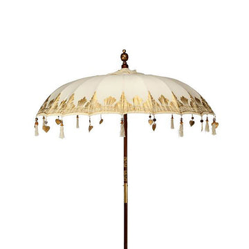 Sombrilla de patio estilo Bali en lona color crema con detalles dorados, flecos y poste de madera sobre fondo blanco